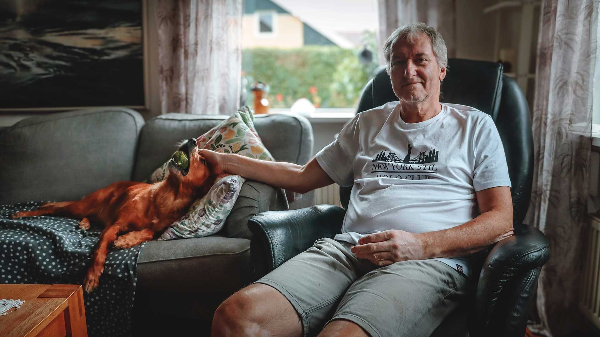 Per-Håkan in an armchair in his living room, with a red-haired dog stretched out on the sofa beside him