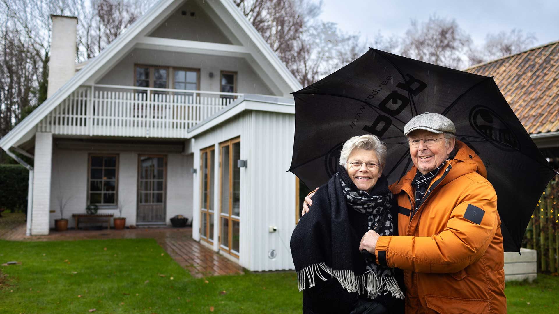 Stellan and Monica Lindsjö under a black umbrella in front of their white house in Trelleborg